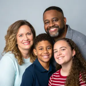 Four people, two adults and two children, pose closely together in front of a plain gray background, all smiling at the camera.