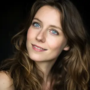 A woman with long wavy brown hair and blue eyes smiles softly against a dark background.