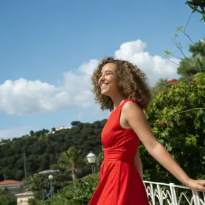 A woman in a red dress stands outdoors, smiling and leaning on a white railing, with green trees and a blue sky with clouds in the background.