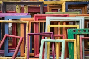 A variety of empty, colorful wooden picture frames of different sizes are stacked and overlapping in a random arrangement.