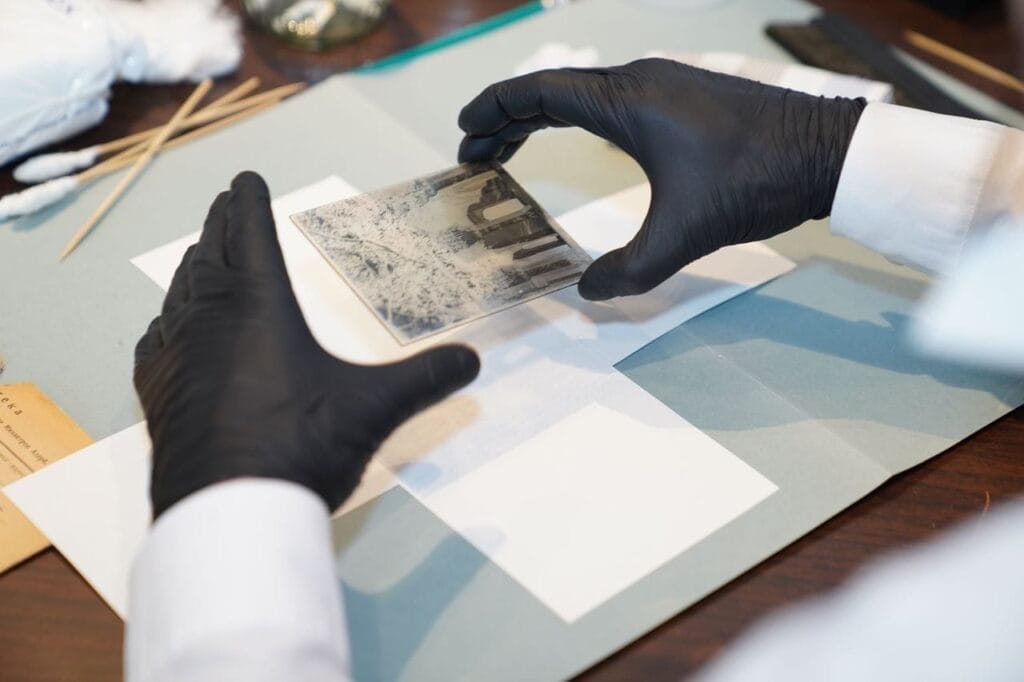 Person wearing black gloves handles a photographic glass plate negative over a work surface with white paper and tools.