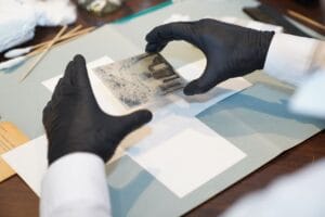 Person wearing black gloves handles a photographic glass plate negative over a work surface with white paper and tools.