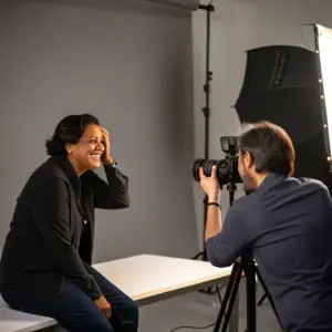 A person sits and smiles while another person takes their photo in a professional studio with lighting equipment in the background.