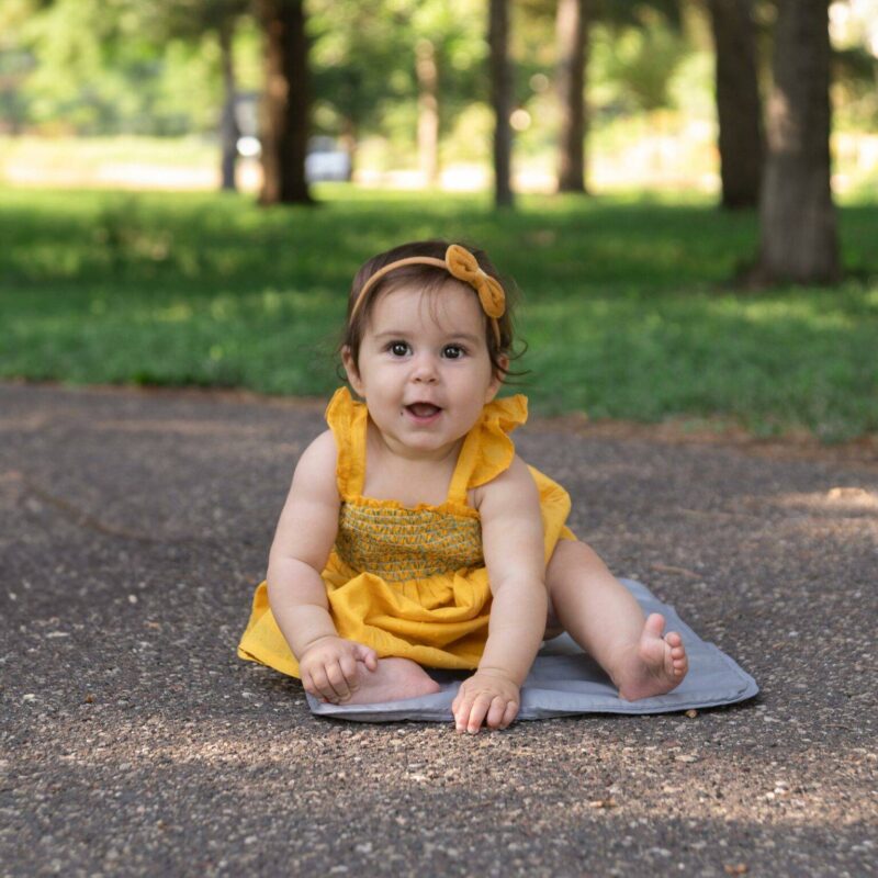 A baby in a yellow dress sits on a gray mat on a paved path in the park, surrounded by green trees, creating an enchanting scene of children photography.
