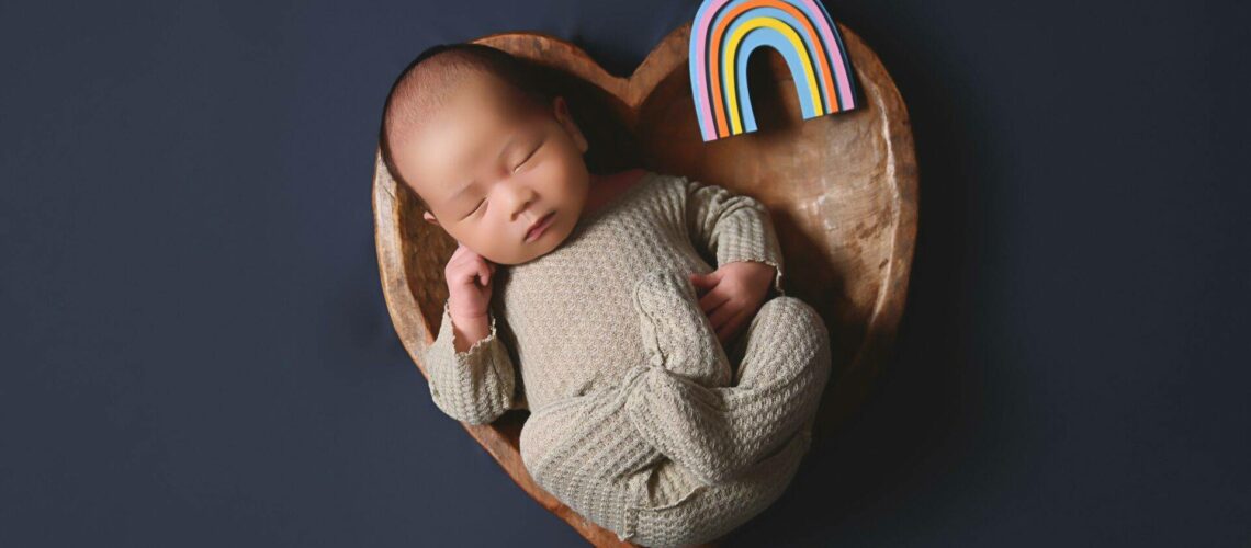 A baby sleeps in a heart-shaped wooden bowl with a small rainbow figure beside them on a dark background.