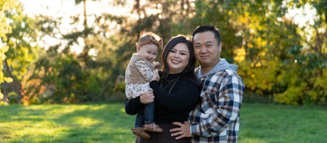 A family of three poses outdoors, with the woman holding a child, and the man standing to her right. Trees and grass fill the background.