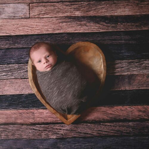 A baby boy in a heart shaped bowl on a wooden floor.