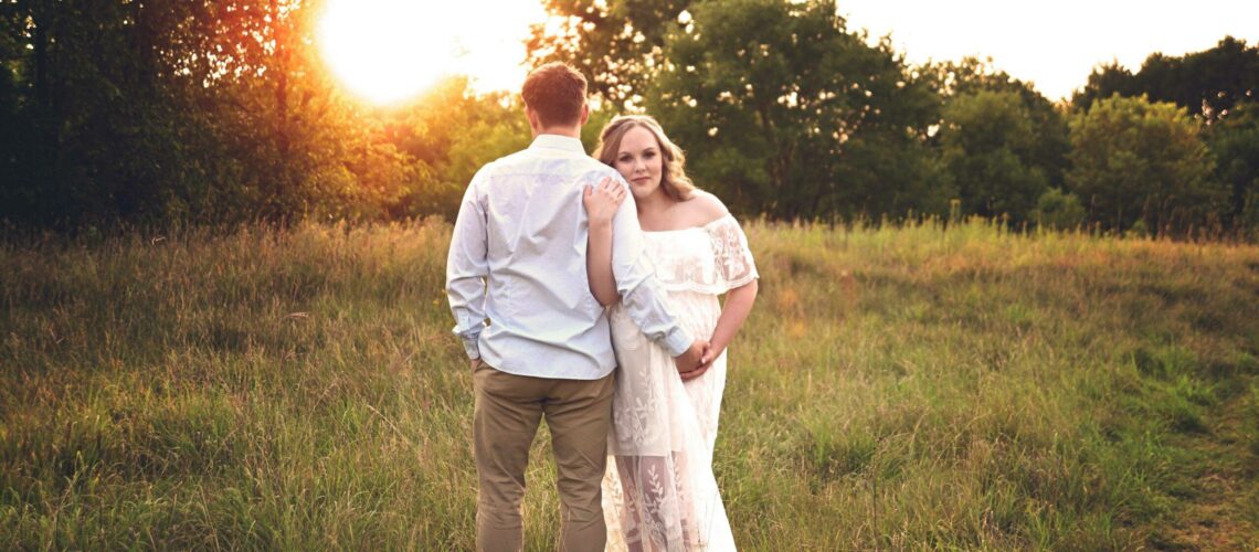 A pregnant woman and her husband in a field light by the golden hour sun.