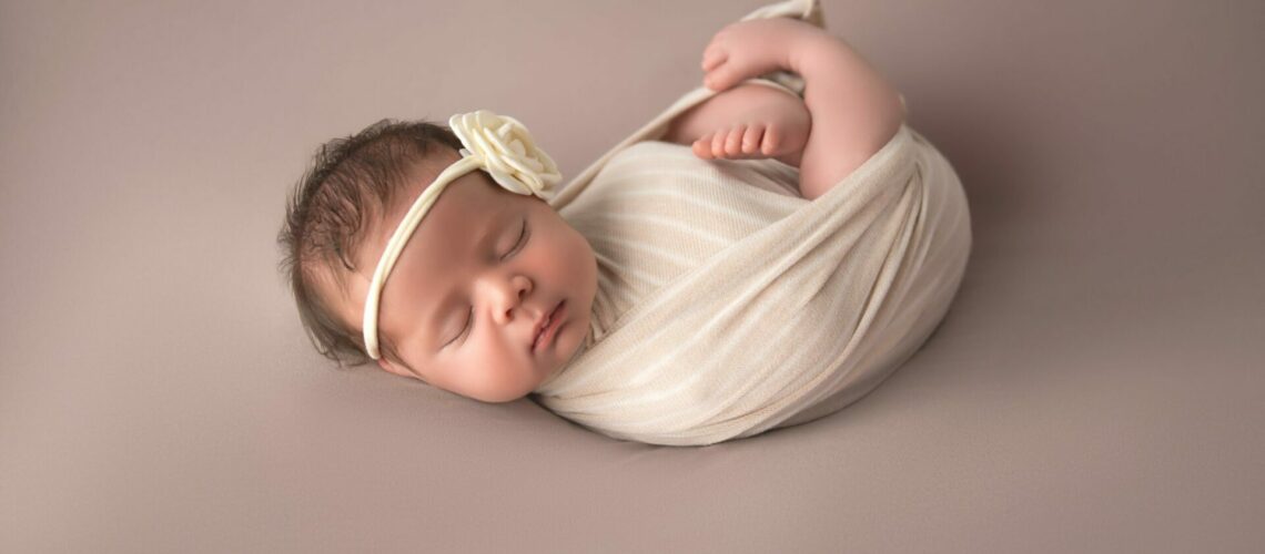 A newborn girl wrapped in a white blanket on a gray background.