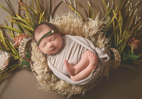 A baby girl is laying in a basket with grass and flowers.