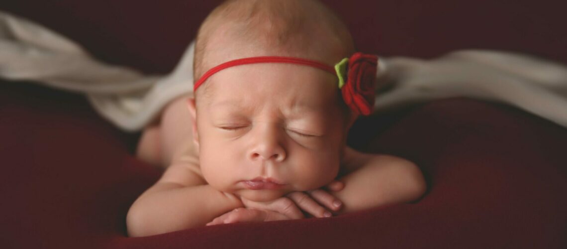 A baby girl wearing a red flower headband is laying on a red blanket.