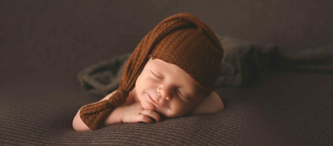 A baby wearing a brown knitted hat is laying on a blanket.