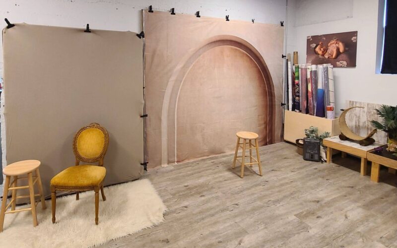 Photography studio setup with a beige backdrop, two stools, a vintage chair on a rug, and photography equipment including rolls of background paper and lighting accessories.