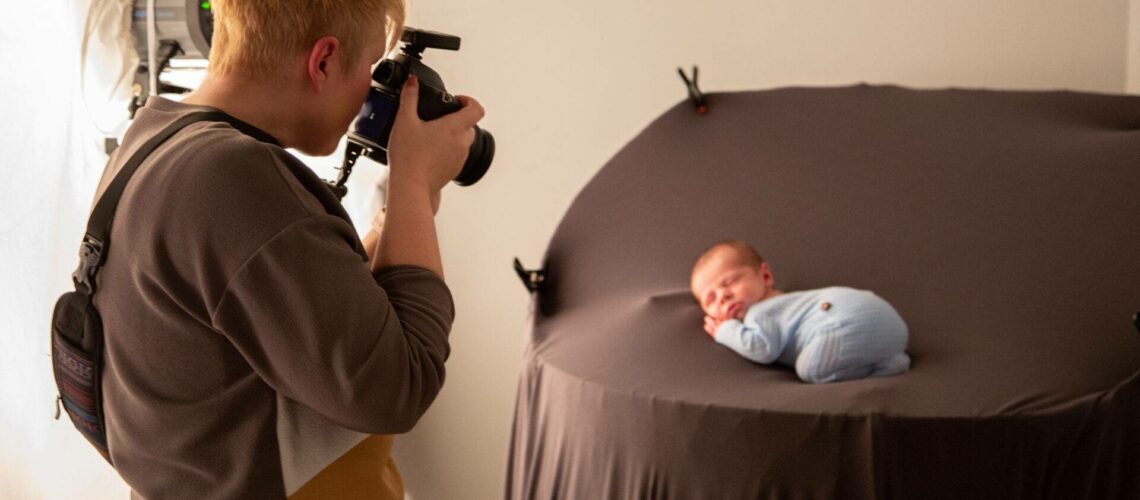 A photographer with a camera captures a photo of a newborn baby swaddled in blue, lying on a brown circular backdrop.