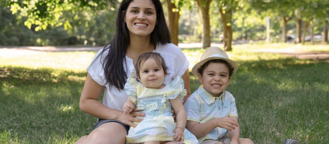 A woman sits on grass with two young children, a toddler and a slightly older boy in a hat, all smiling in a park setting with trees in the background.