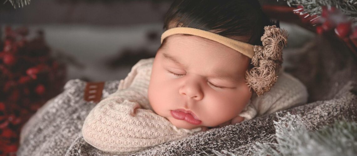 A sleeping baby with a headband is nestled in a cozy, textured blanket surrounded by pine branches.