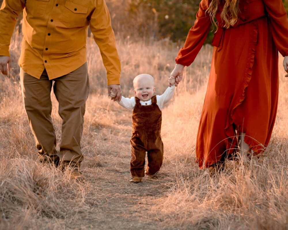 A small child walks through a dry grassy field holding hands with two adults, all dressed in autumn-colored clothing.