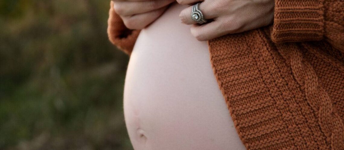 A pregnant person stands outdoors, gently holding dried flowers. They wear a brown knit cardigan and jeans, with a visible pregnant belly.