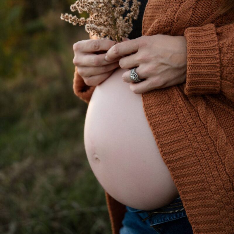 A pregnant person stands outdoors, gently holding dried flowers. They wear a brown knit cardigan and jeans, with a visible pregnant belly.