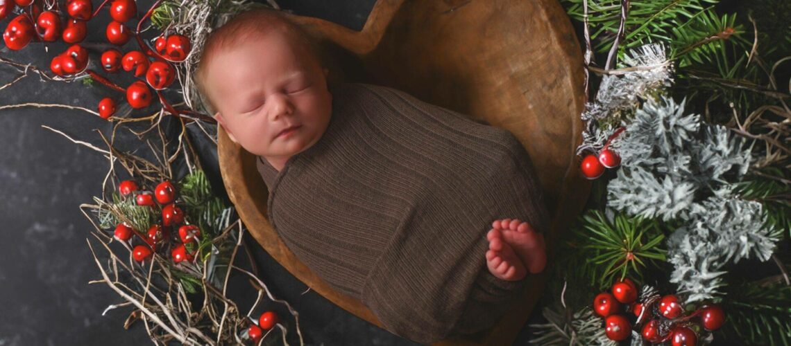 A newborn baby wrapped in brown fabric sleeps in a heart-shaped wooden bowl, surrounded by greenery and red berries on a dark background.