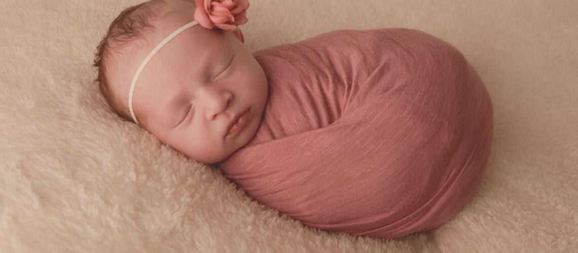 A newborn girl wrapped in a pink wrap sleeping on a fluffy blanket.