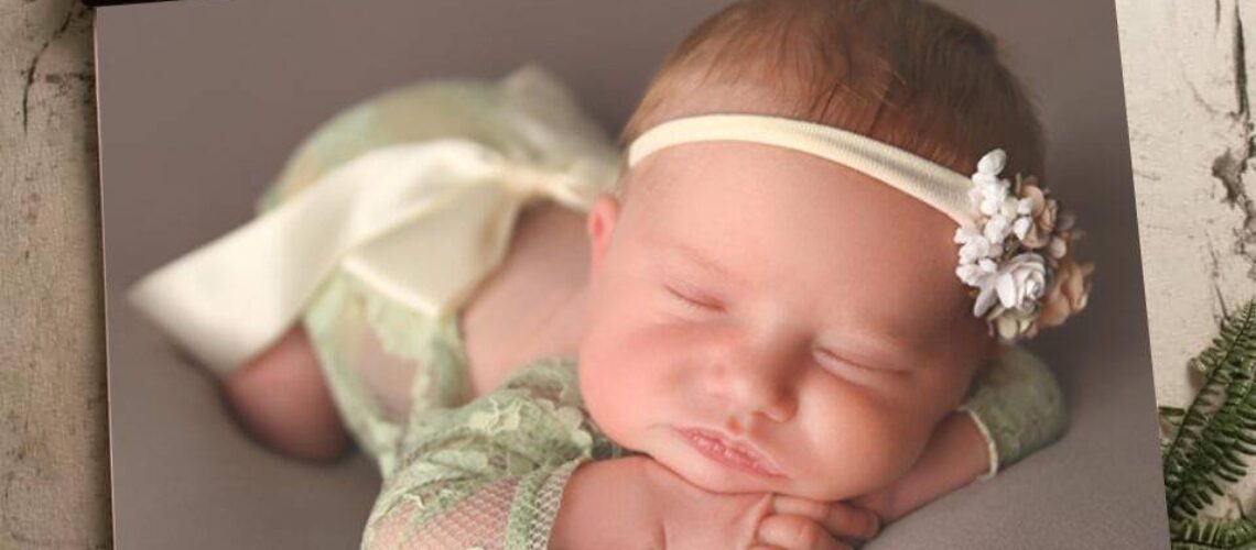 A baby in a green lace outfit and headband sleeps on a wooden frame background.