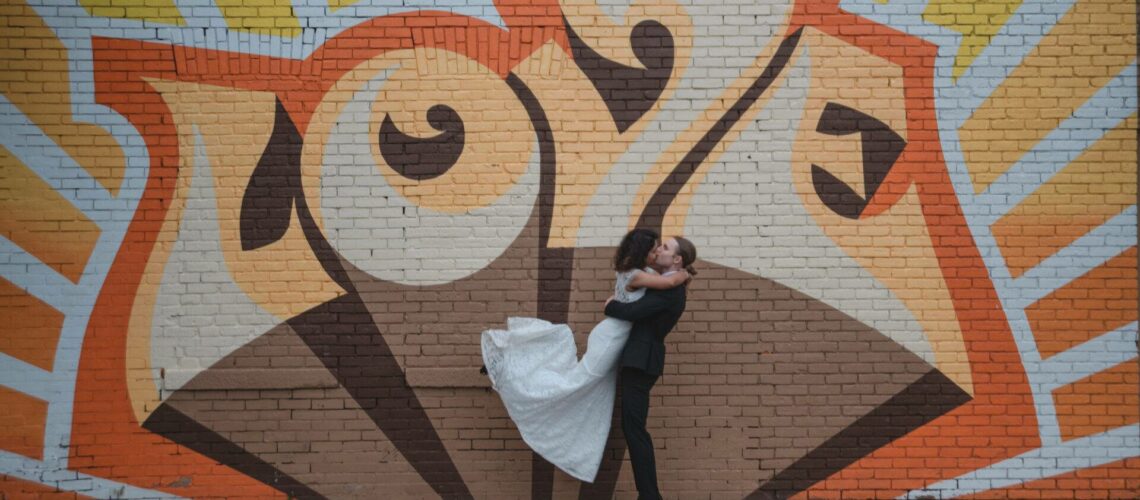 A couple embraces and kisses in front of a colorful mural with the word "love" on a brick wall. The background features orange and yellow rays.