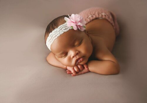 A baby girl is laying down with a pink flower headband on her head.