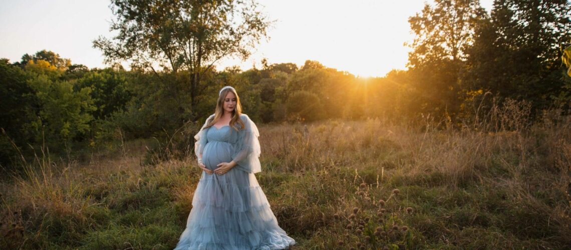 A pregnant woman in a flowing gown stands in a field at sunset, surrounded by trees and tall grass.