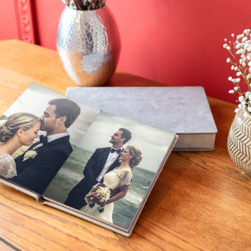 An open photo album on a wooden table shows a couple in wedding attire. A vase with white flowers and a silver decorative item are in the background against a red wall.