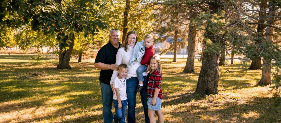 A family of five poses together outdoors in a wooded area, standing on grass with trees and sunlight in the background.