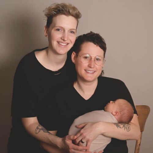 Two smiling individuals with short hair sit together. One holds a sleeping baby wrapped in a beige blanket. Both wear black shirts and have visible tattoos on their arms.