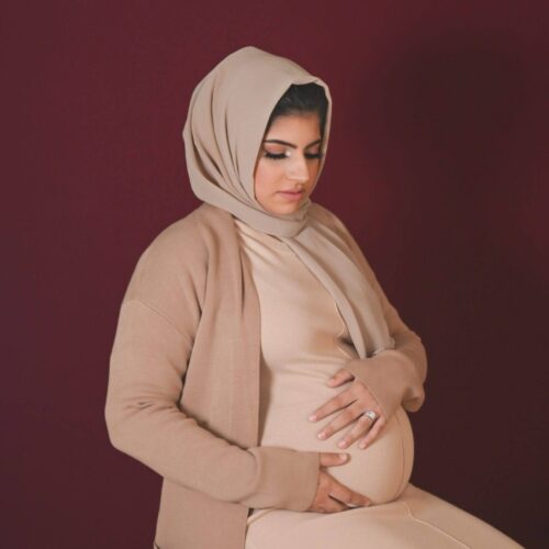 A pregnant woman wearing a beige hijab and matching outfit sits on a stool, holding her baby bump against a maroon background.