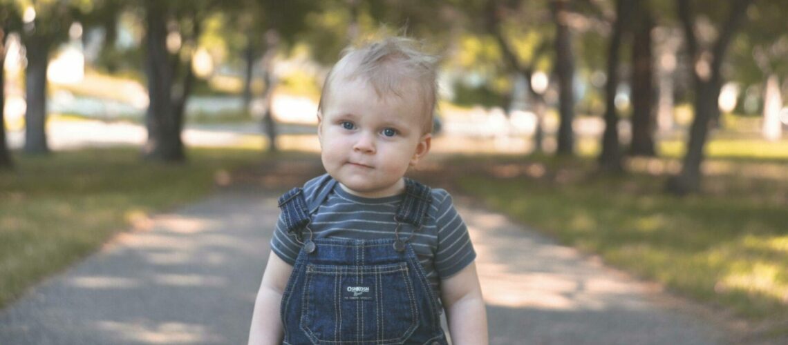 A baby boy in overalls standing on a path.