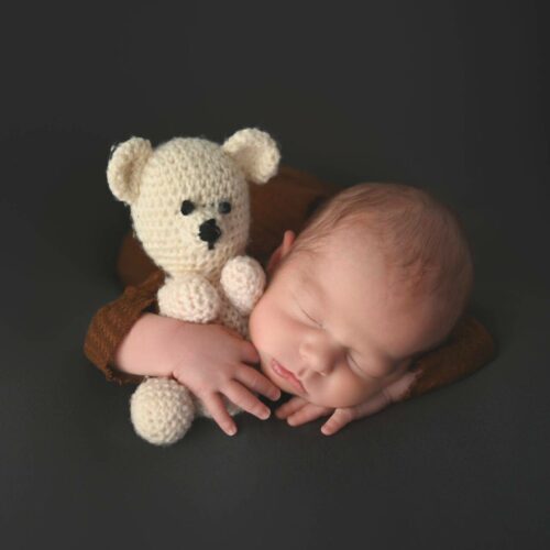 A sleeping newborn baby cradles a small, knitted bear on a dark surface.