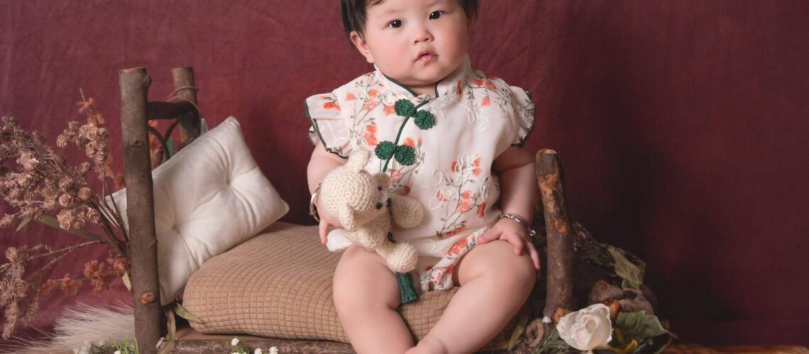 A toddler in a floral outfit sits on a small wooden bench, holding a stuffed toy, surrounded by flowers and cushions against a maroon backdrop.