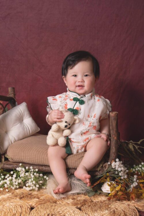 A baby sits on a rustic wooden bench, holding a small stuffed bear, surrounded by flowers and cushions, against a maroon backdrop.