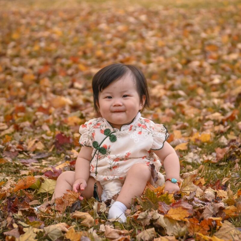 A baby in a floral outfit sits on a lawn covered with autumn leaves, surrounded by scattered red, yellow, and brown foliage.