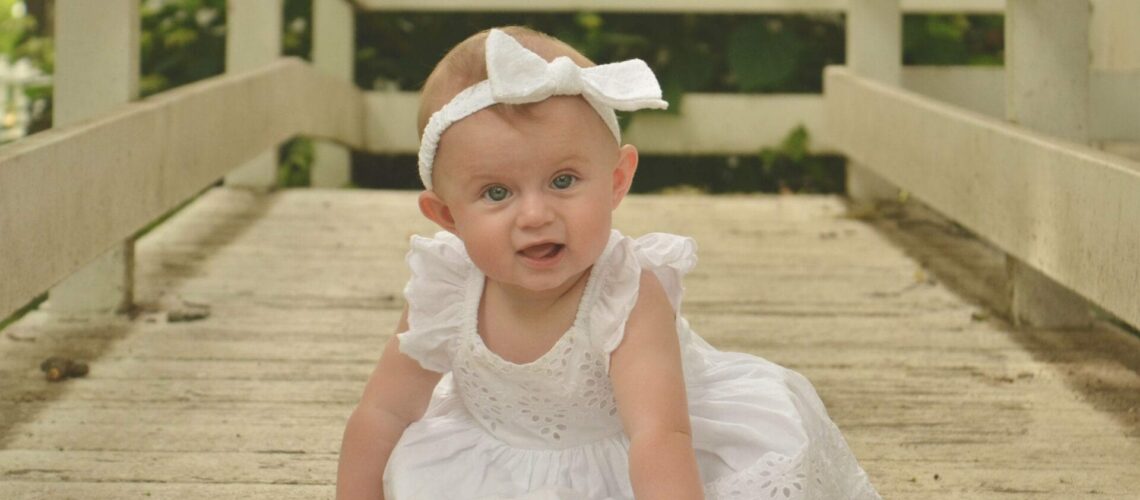 A baby girl in a white dress sitting on a wooden bridge.
