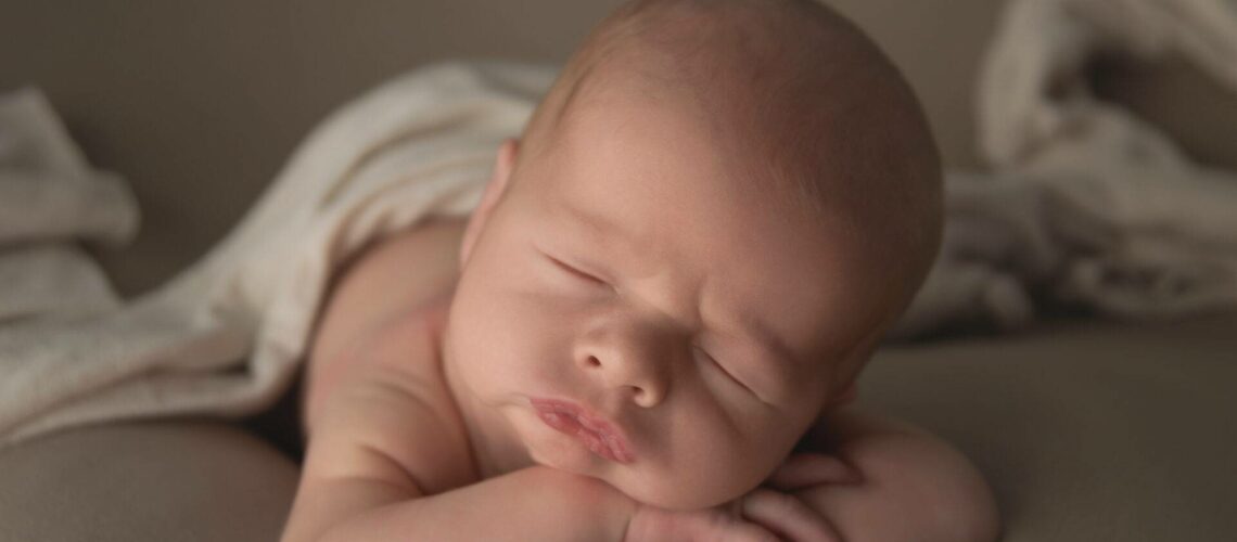 A newborn baby sleeps on its stomach with a blanket partially covering its body. The baby has its head resting on its crossed arms.