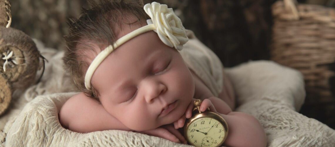 Sleeping baby with a headband and flower, lying on a soft blanket in a wicker basket, holding an antique pocket watch.