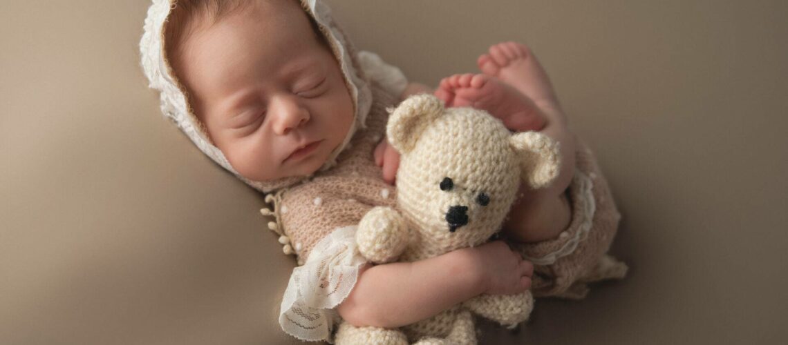 A newborn baby sleeps peacefully while wearing a white knit bonnet and holding a crocheted teddy bear. The baby has eyes closed and is wrapped in a neutral-colored blanket.