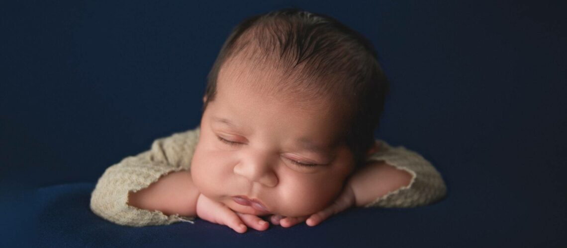 Newborn baby peacefully sleeping with head resting on hands against a dark blue background.