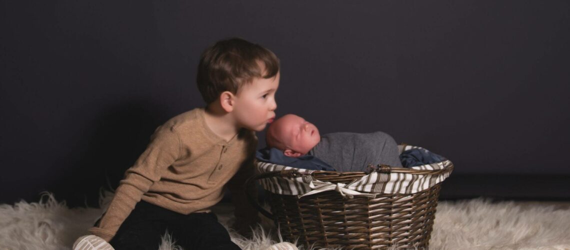 A young child sitting on a white rug leans towards a newborn baby wrapped in a gray blanket, resting in a wicker basket. The background is dark, highlighting the children.