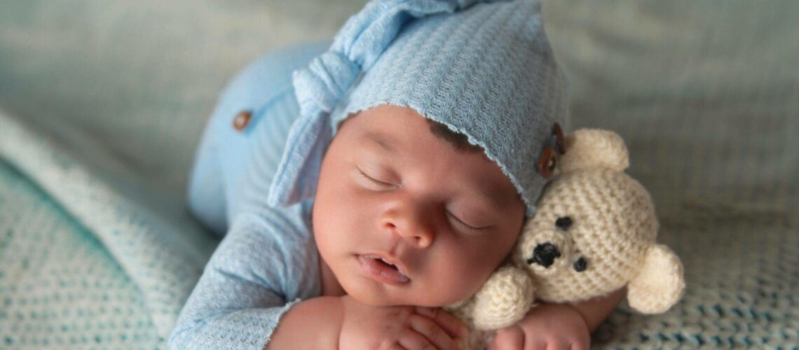 A sleeping baby in blue pajamas and hat, resting on a knitted blanket, cuddles a small crocheted teddy bear.