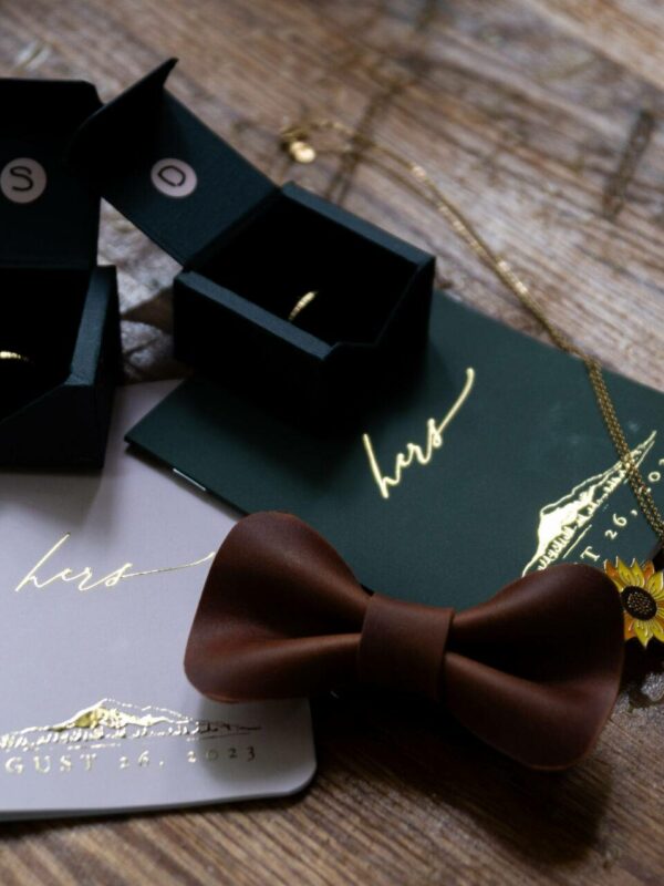 Wooden table with wedding items: ring boxes, vows booklets labeled "hers", a brown bow tie, and gold jewelry including a sunflower pendant.