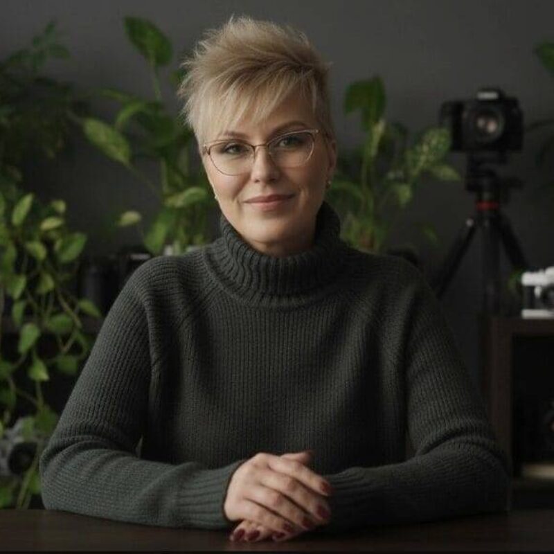 A person with short blond hair and glasses sits at a table, wearing a dark sweater, with cameras and green plants in the background.