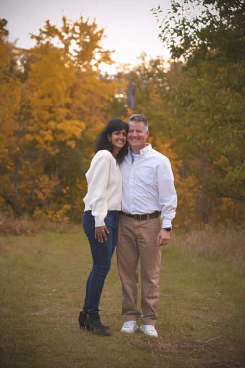 A woman and a man stand close together outdoors on a grassy path, surrounded by trees with autumn leaves.