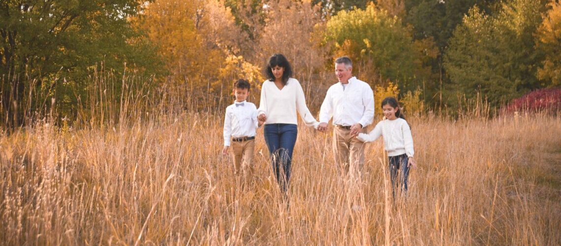 A family of four walks through a grassy field with autumn trees in the background, all wearing light-colored tops and tan pants.