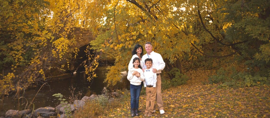 A family of four stands together by a creek in a park, surrounded by trees with yellow autumn leaves.